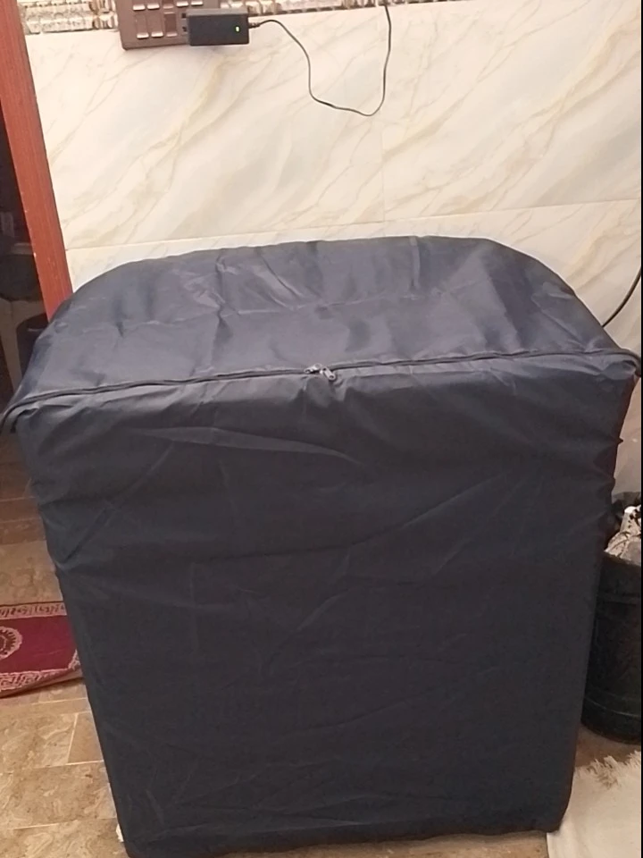 Dark grey dustproof washing machine cover with top zipper access fitted over a washing machine placed against a tiled wall.